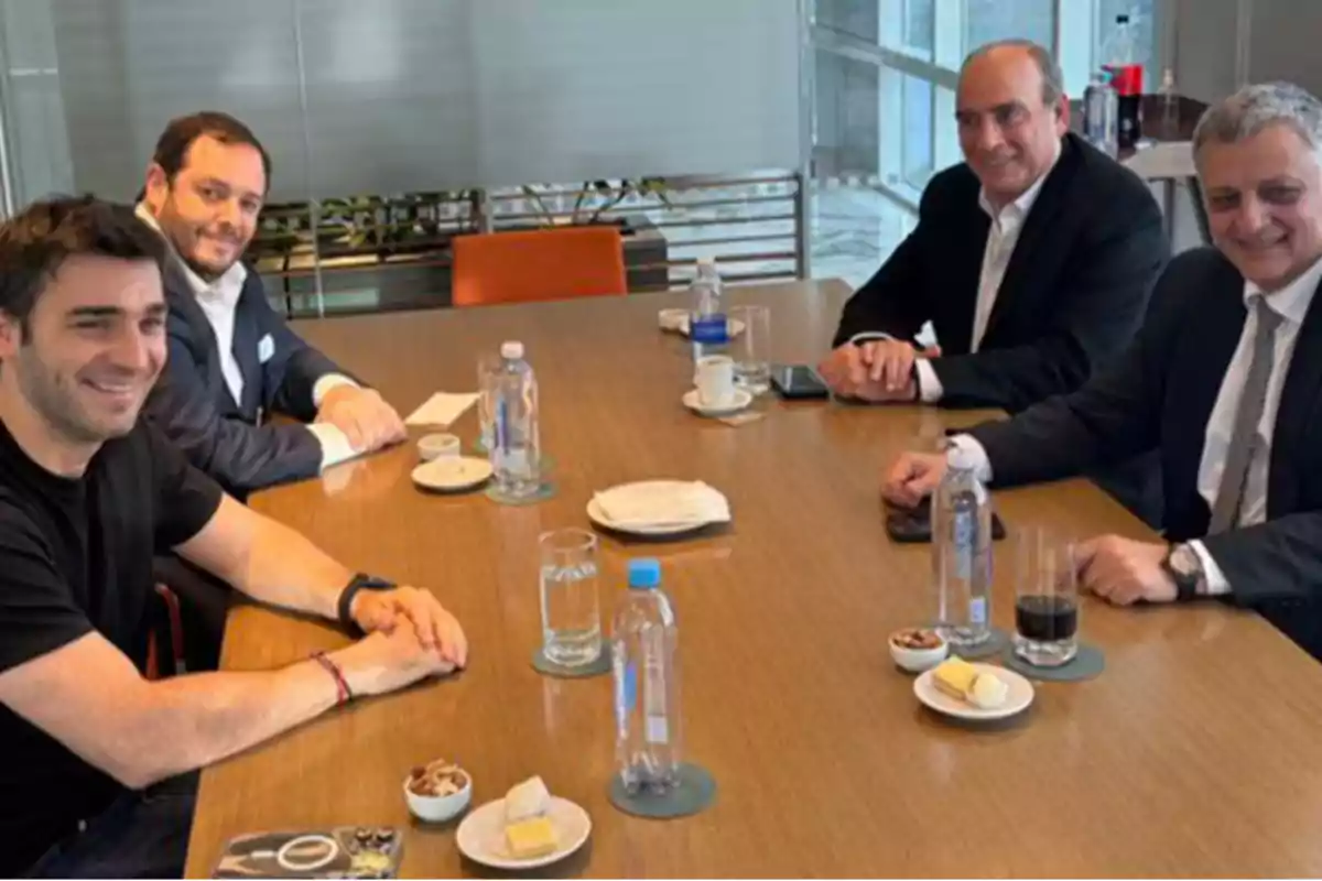 Four men sitting around a wooden table in a meeting room, with water bottles, glasses, coffee, and small plates with snacks. Four men sitting around a wooden table in a meeting room, with water bottles, glasses, coffee, and small plates with snacks.