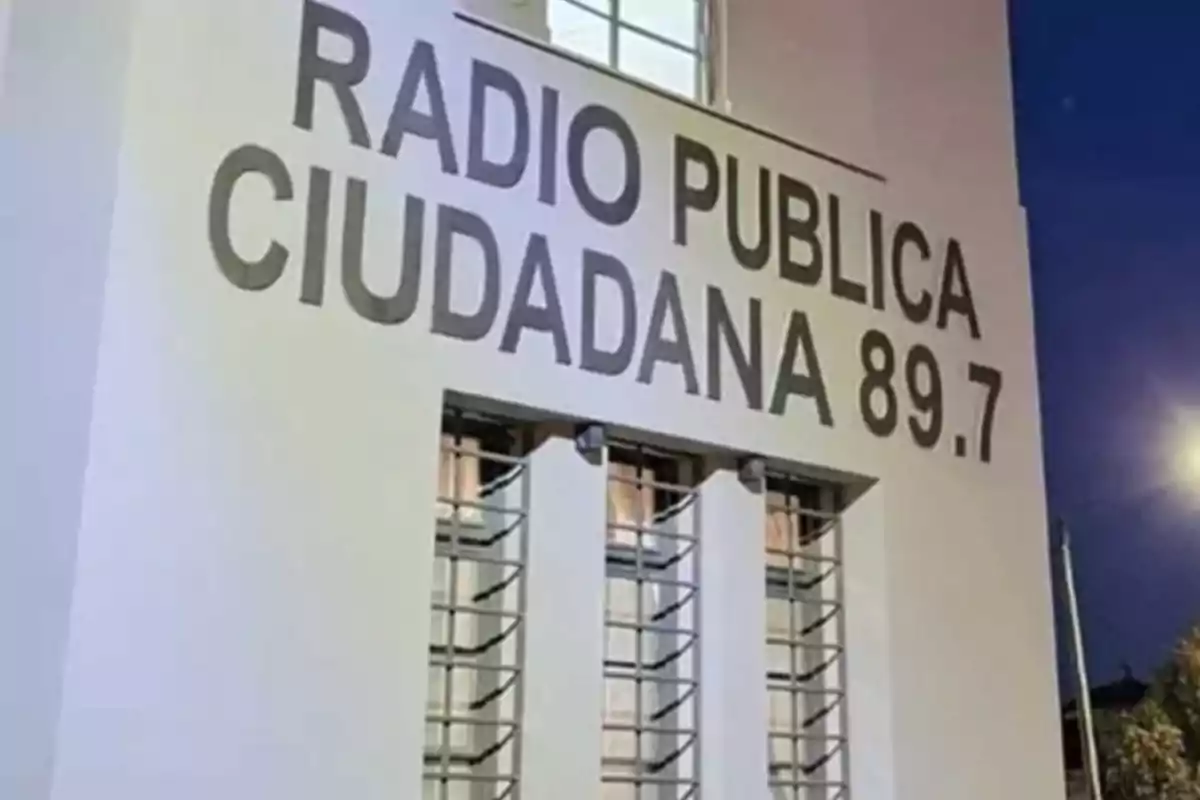 Edificio blanco con letras grandes que dicen RADIO PUBLICA CIUDADANA 89.7 y ventanas con rejas verticales