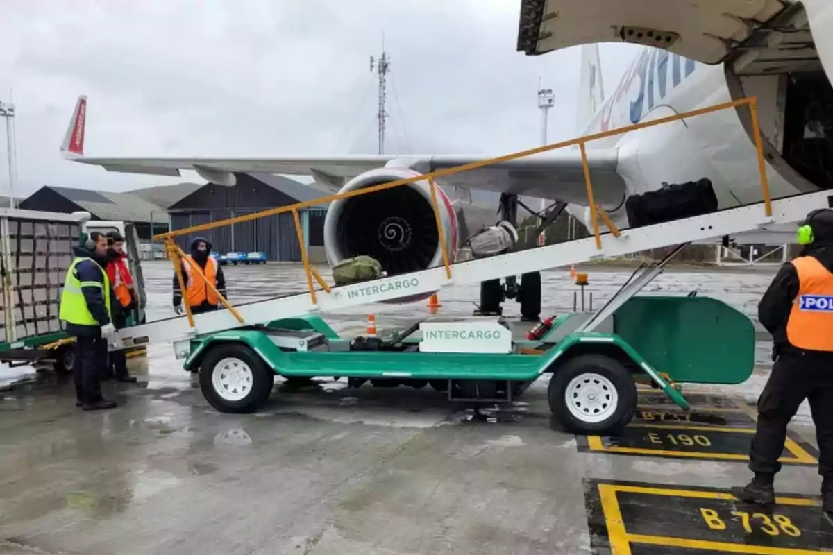 Trabajadores con chalecos de seguridad cargan equipaje en la bodega de un avión en una pista mojada usando una cinta transportadora