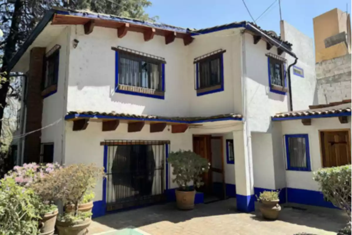 Two-story house with white walls and blue details, barred windows, and several potted plants at the entrance. Two-story house with white walls and blue details, barred windows, and several potted plants at the entrance.