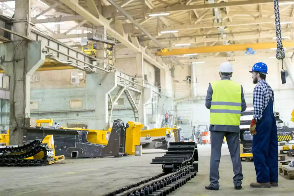 Dos trabajadores con casco y chaleco observan maquinaria pesada dentro de una fábrica industrial Dos trabajadores con casco y chaleco observan maquinaria pesada dentro de una fábrica industrial