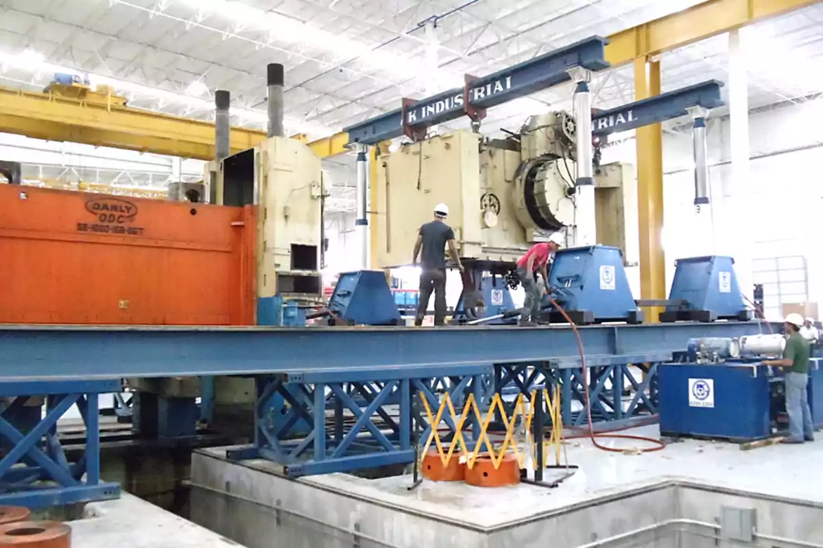 Trabajadores con casco operan maquinaria industrial pesada en el interior de una fábrica con estructura metálica azul y amarilla Trabajadores con casco operan maquinaria industrial pesada en el interior de una fábrica con estructura metálica azul y amarilla