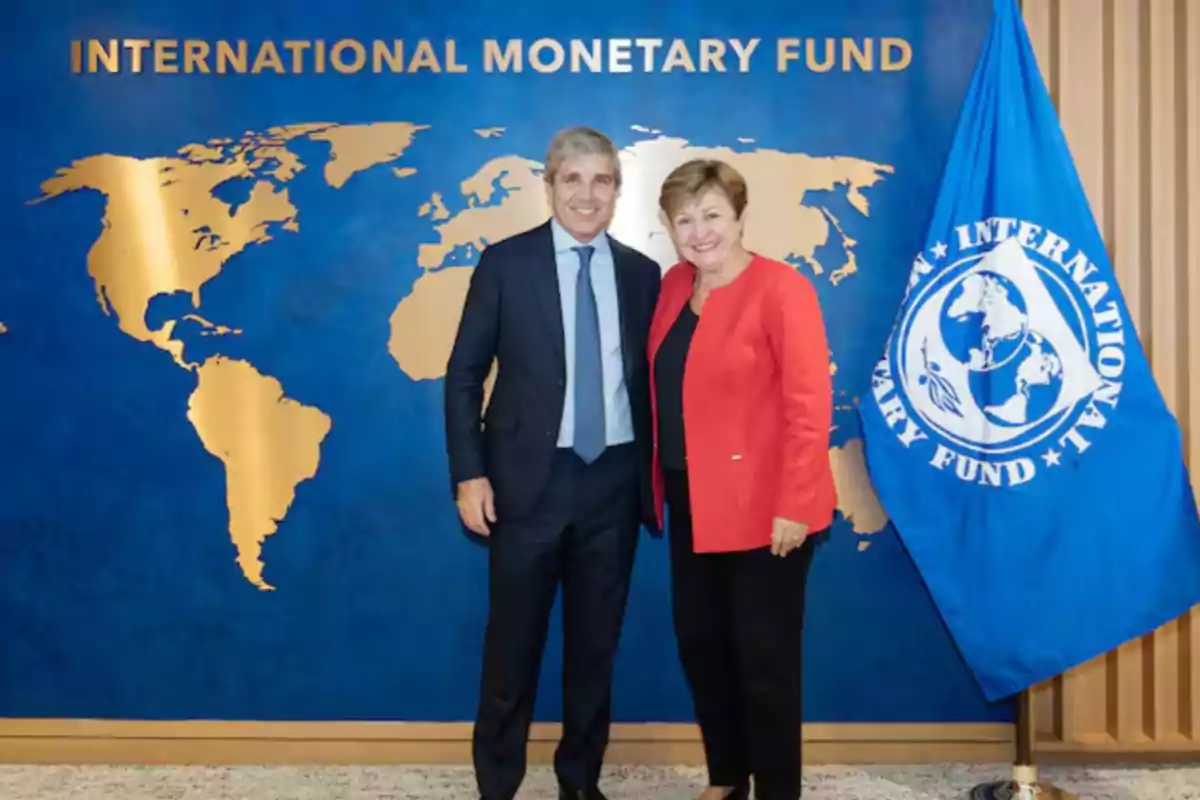 Dos personas posan sonrientes frente a un fondo azul con un mapa dorado del mundo y el texto International Monetary Fund junto a una bandera azul del Fondo Monetario Internacional Dos personas posan sonrientes frente a un fondo azul con un mapa dorado del mundo y el texto International Monetary Fund junto a una bandera azul del Fondo Monetario Internacional
