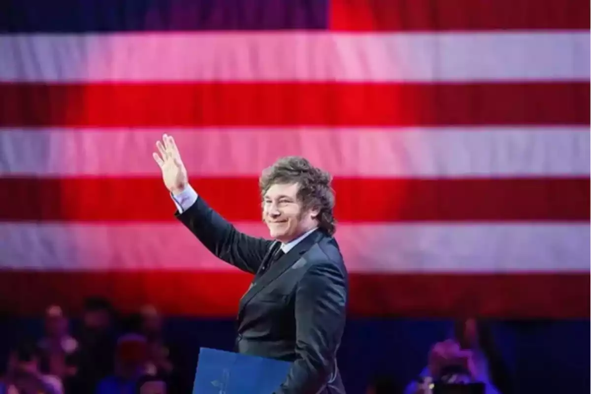 Hombre de traje saludando con la mano levantada frente a una gran bandera de Estados Unidos Hombre de traje saludando con la mano levantada frente a una gran bandera de Estados Unidos