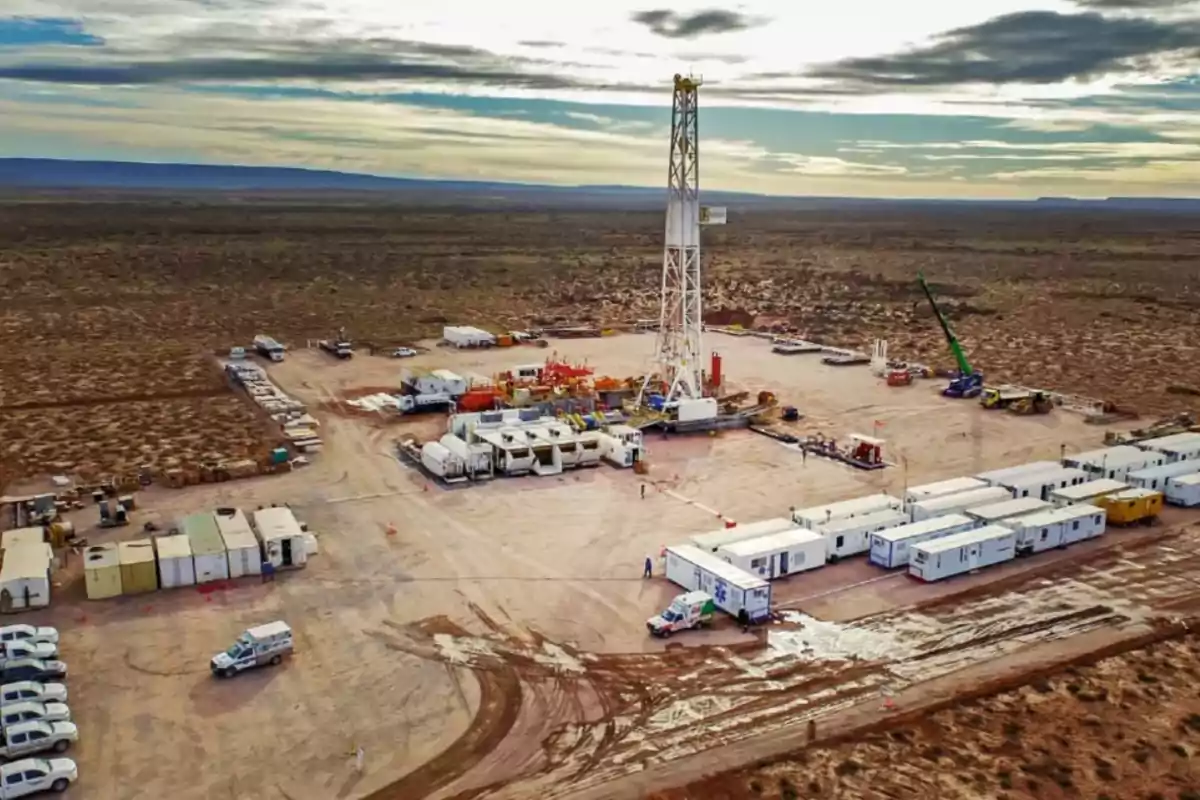 Vista aérea de un campo petrolero con una torre de perforación rodeada de vehículos, módulos y maquinaria en una zona desértica bajo un cielo parcialmente nublado Vista aérea de un campo petrolero con una torre de perforación rodeada de vehículos, módulos y maquinaria en una zona desértica bajo un cielo parcialmente nublado