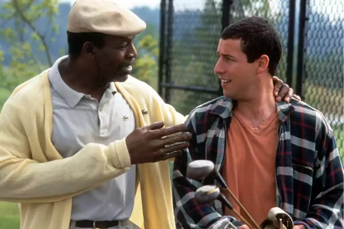 Two men are talking on a golf course; one of them is wearing a beret and a yellow sweater, while the other is holding a bag of golf clubs and wearing a plaid jersey.