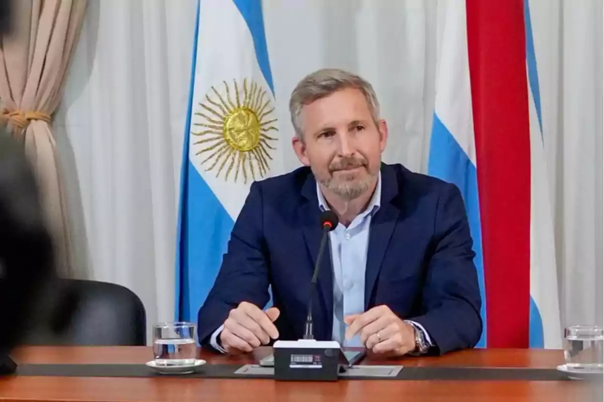 Hombre de cabello canoso y barba sentado en una mesa con micrófono y vasos de agua, al fondo banderas de Argentina y otra bandera tricolor