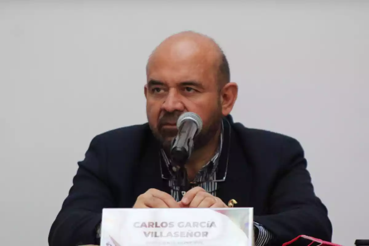 Hombre calvo con barba sentado frente a un micrófono y una placa con su nombre en una mesa durante una conferencia