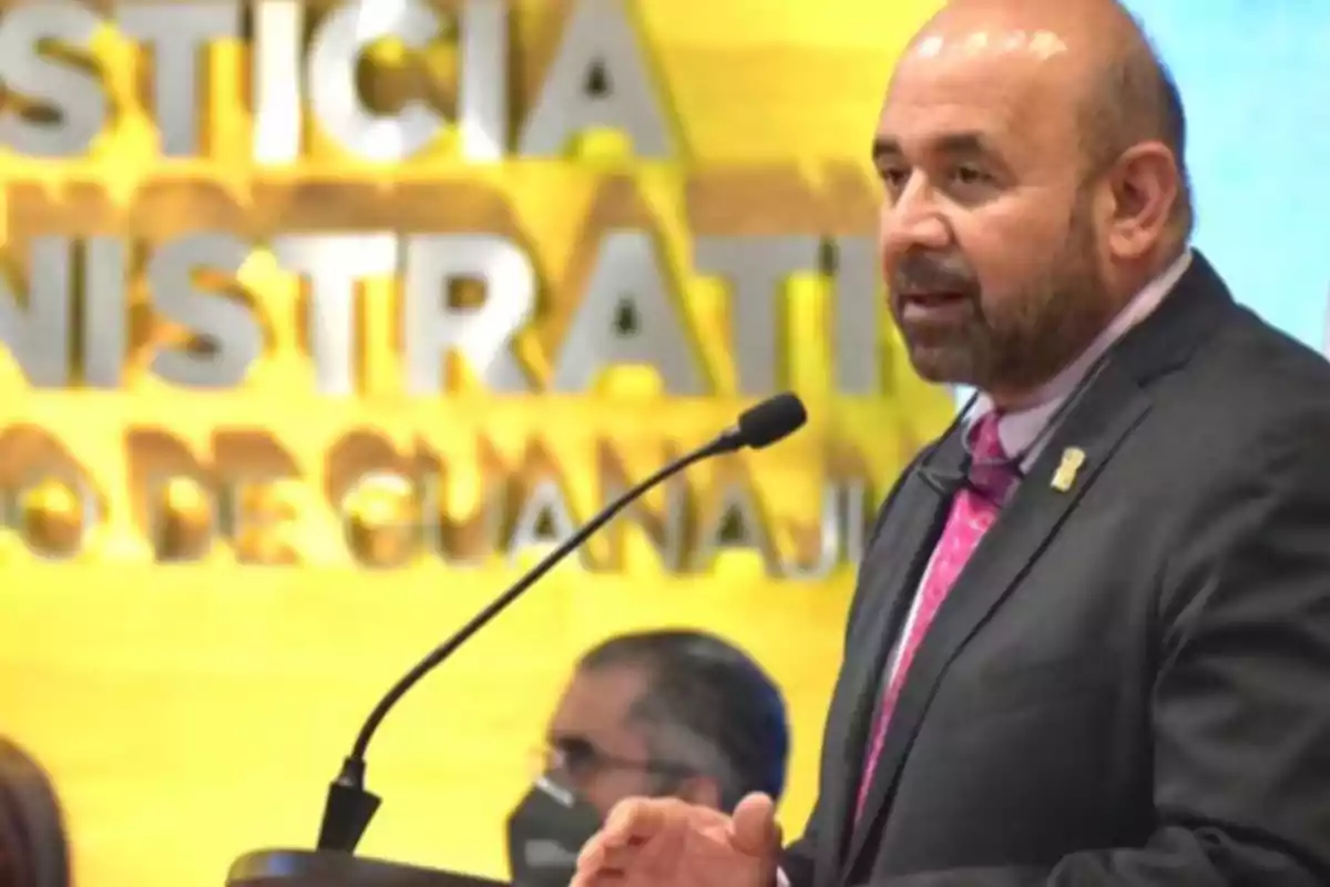 Hombre con barba y traje oscuro hablando en un micrófono frente a un fondo amarillo con letras grandes desenfocadas Hombre con barba y traje oscuro hablando en un micrófono frente a un fondo amarillo con letras grandes desenfocadas