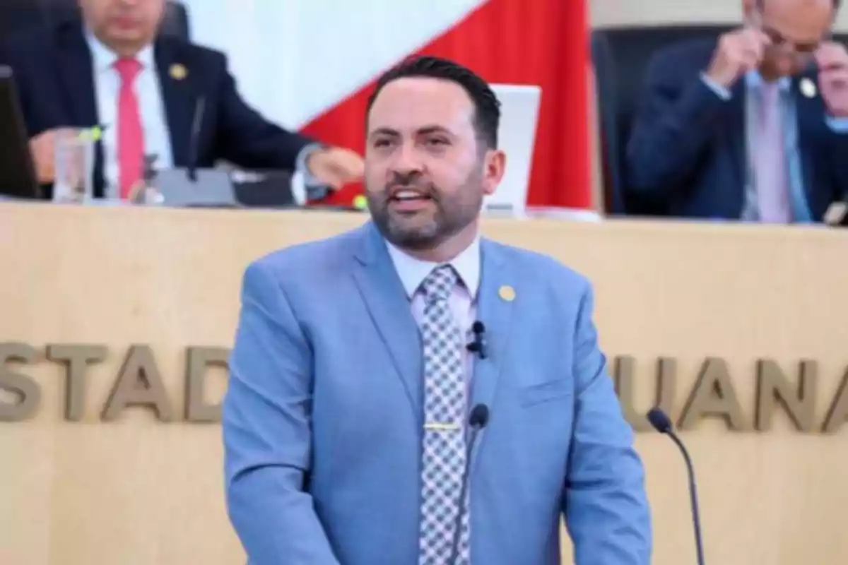 Hombre con saco azul claro hablando en un podio en un recinto oficial con personas al fondo Hombre con saco azul claro hablando en un podio en un recinto oficial con personas al fondo