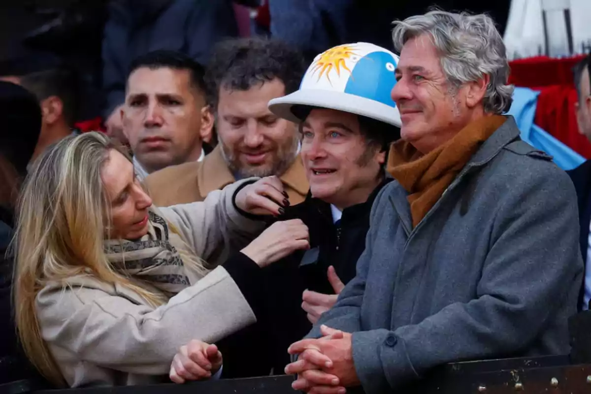 Un grupo de personas sonríe y conversa en un evento al aire libre, una mujer ajusta la ropa de un hombre que lleva un casco decorado con los colores de la bandera argentina y un sol, todos visten ropa de abrigo Un grupo de personas sonríe y conversa en un evento al aire libre, una mujer ajusta la ropa de un hombre que lleva un casco decorado con los colores de la bandera argentina y un sol, todos visten ropa de abrigo