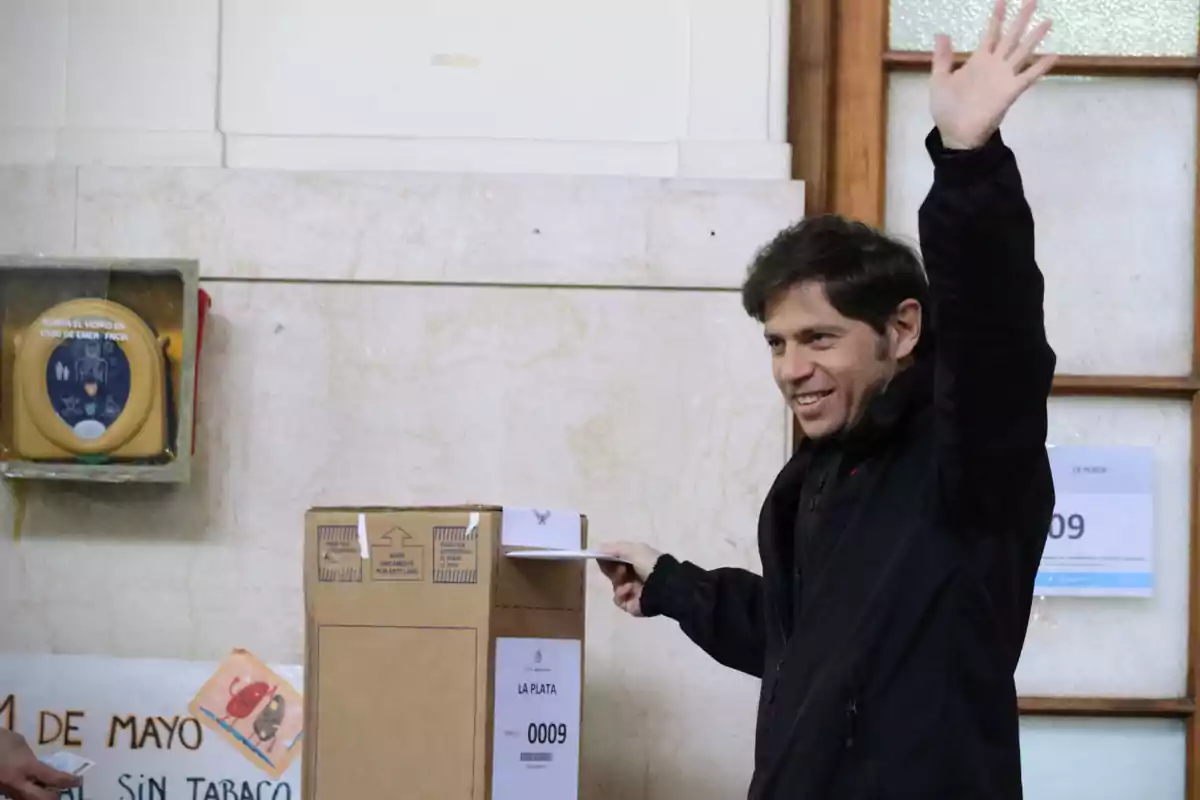 Smiling man in a black jacket voting in a cardboard ballot box and waving with his hand raised in an indoor venue