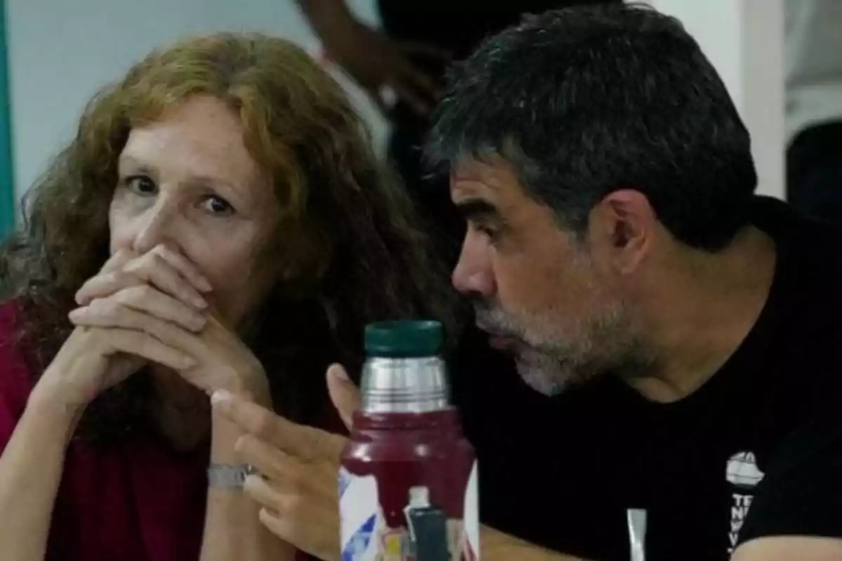 Dos personas sentadas conversando de cerca, una mujer con las manos entrelazadas frente a su rostro y un hombre hablándole al oído, con un termo sobre la mesa