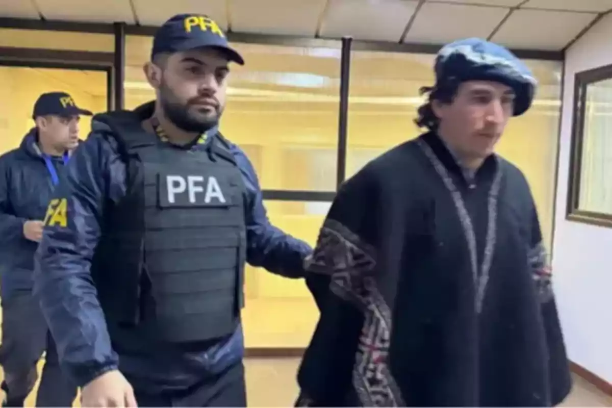 Un hombre con chaleco antibalas y gorra de la PFA escolta a otro hombre con boina y poncho dentro de un edificio.