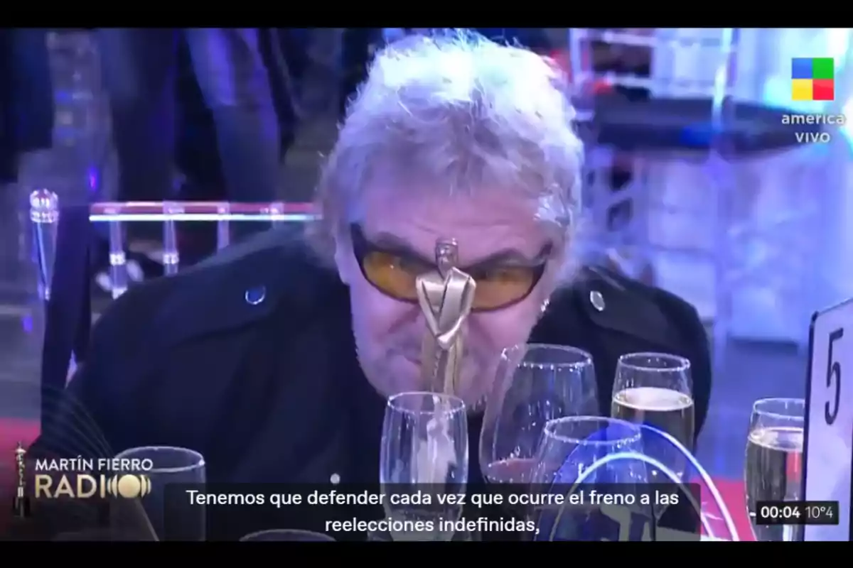 Man with dark glasses sitting at a table during an award ceremony with a golden statuette in front of his face and several glasses around him Man with dark glasses sitting at a table during an award ceremony with a golden statuette in front of his face and several glasses around him