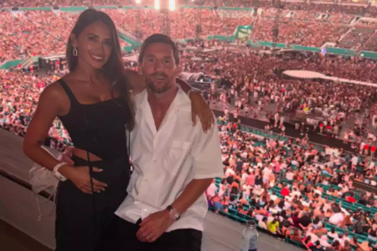 Una pareja sonríe posando en un palco con vista a un estadio lleno de gente durante un concierto. Una pareja sonríe posando en un palco con vista a un estadio lleno de gente durante un concierto.