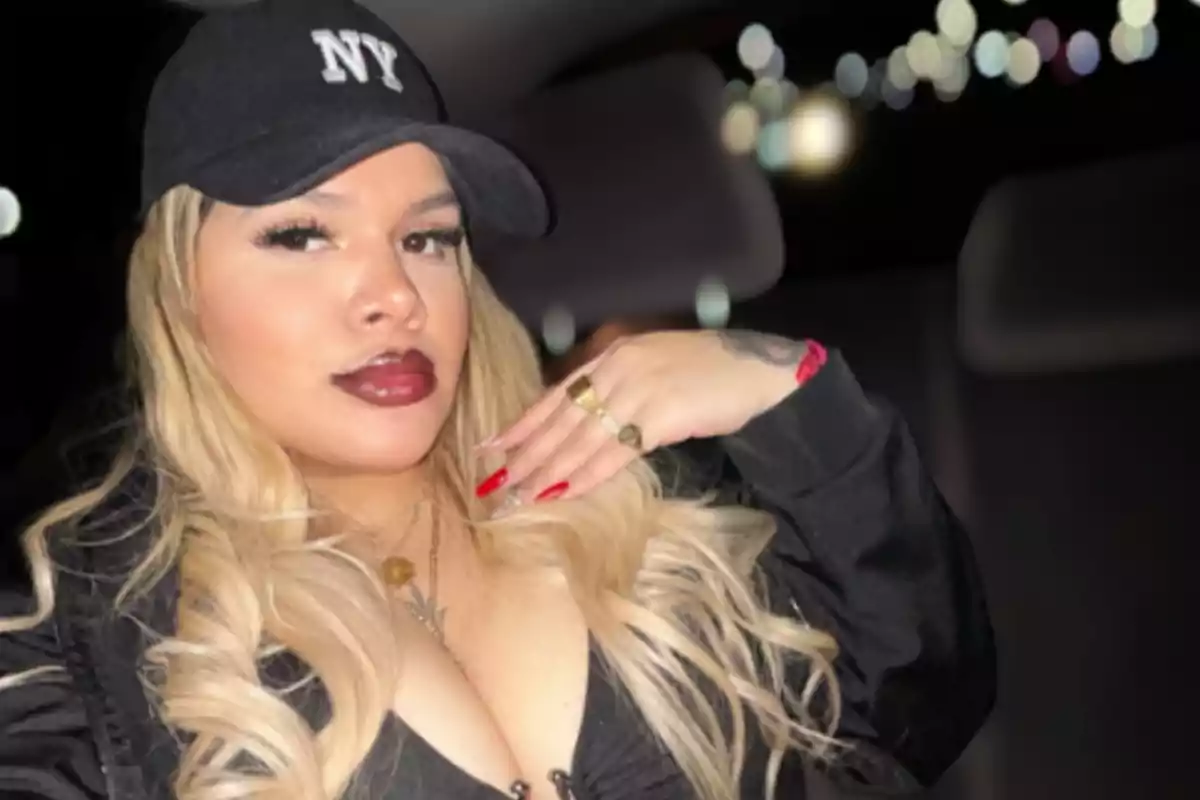 Blonde, long-haired woman wearing a black cap with the letters NY, striking makeup and red nails, sitting inside a car with blurred lights in the background