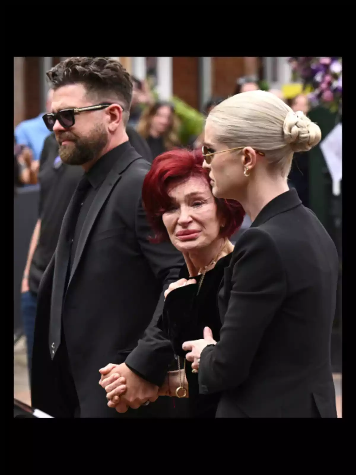 Three people dressed in black, a red-haired woman in the center looks excited and is comforted by a blonde woman while a bearded man with dark glasses stands by her side. Three people dressed in black, a red-haired woman in the center looks excited and is comforted by a blonde woman while a bearded man with dark glasses stands by her side.