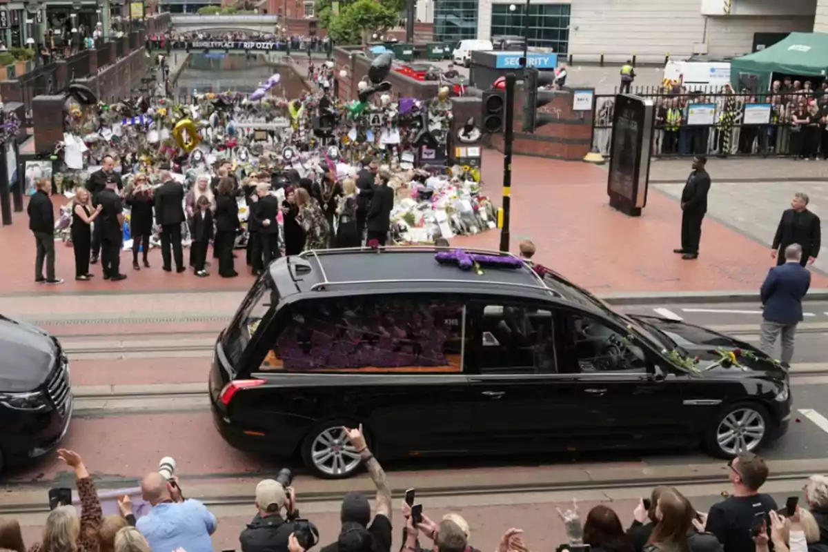 A black hearse moves down a street while a crowd of people dressed in black watch and take photos; in the background, there are flowers and tributes on an urban street corner. A black hearse moves down a street while a crowd of people dressed in black watch and take photos; in the background, there are flowers and tributes on an urban street corner.