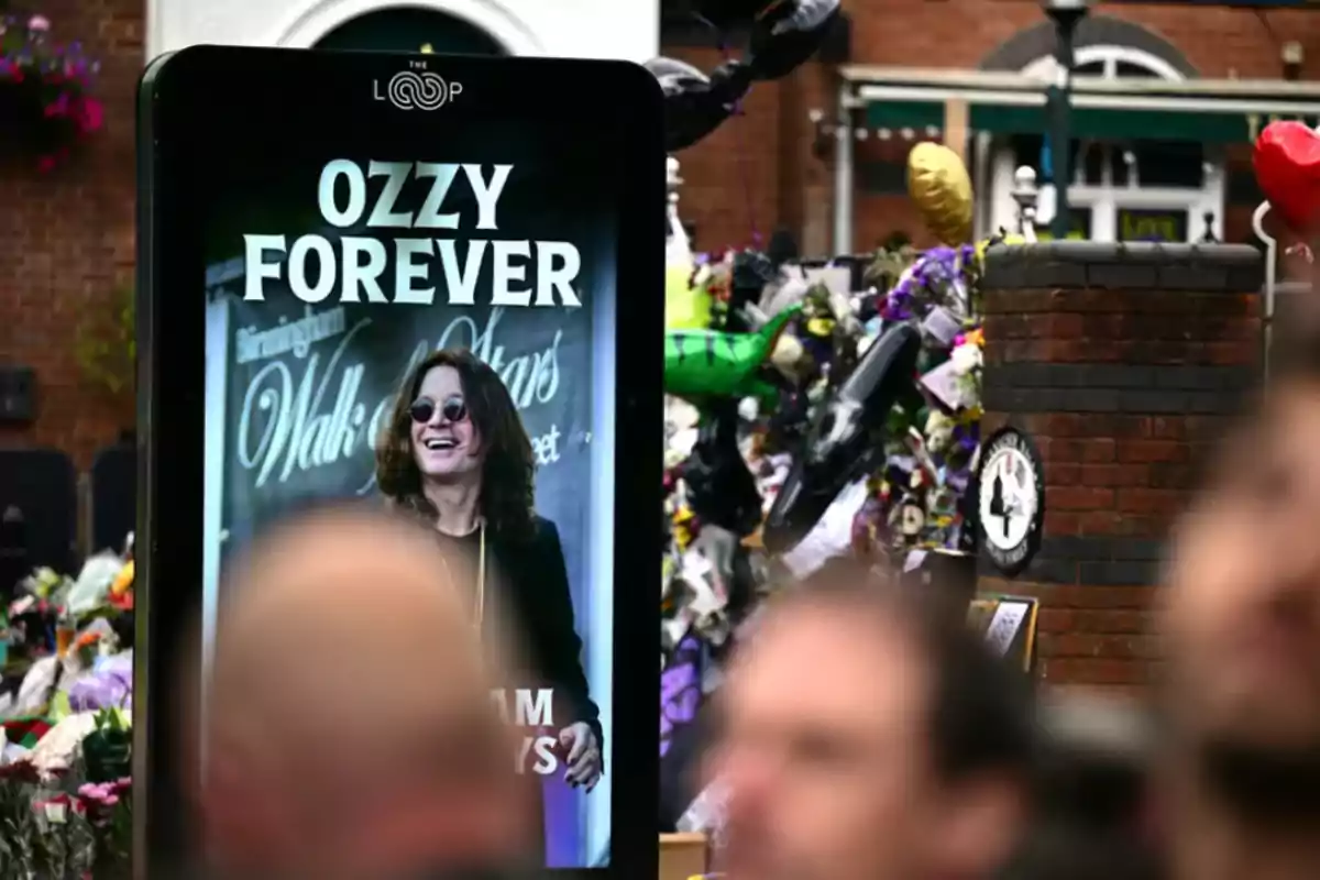 Digital screen with the phrase OZZY FOREVER and the image of a smiling man wearing dark glasses, flowers and balloons in the background on a street with blurred people in the foreground Digital screen with the phrase OZZY FOREVER and the image of a smiling man wearing dark glasses, flowers and balloons in the background on a street with blurred people in the foreground
