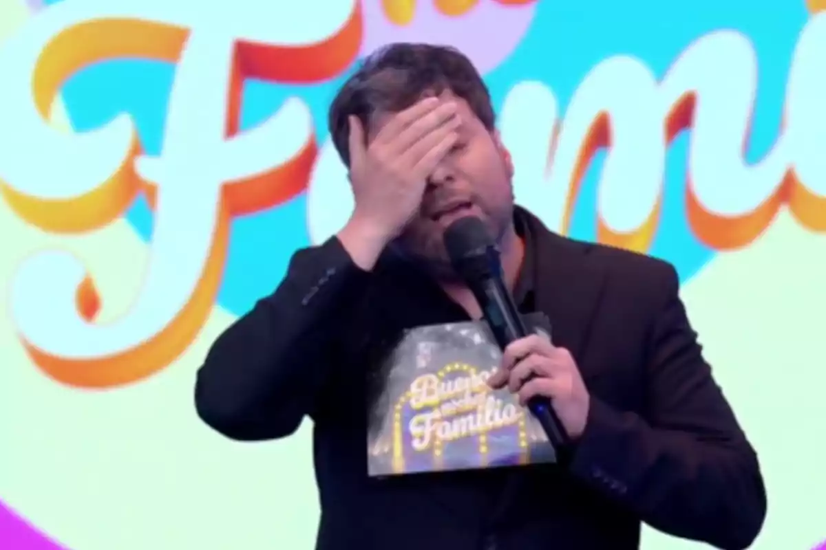 Hombre con barba y saco negro cubriéndose el rostro con una mano mientras sostiene un micrófono y una carpeta en un set colorido de televisión