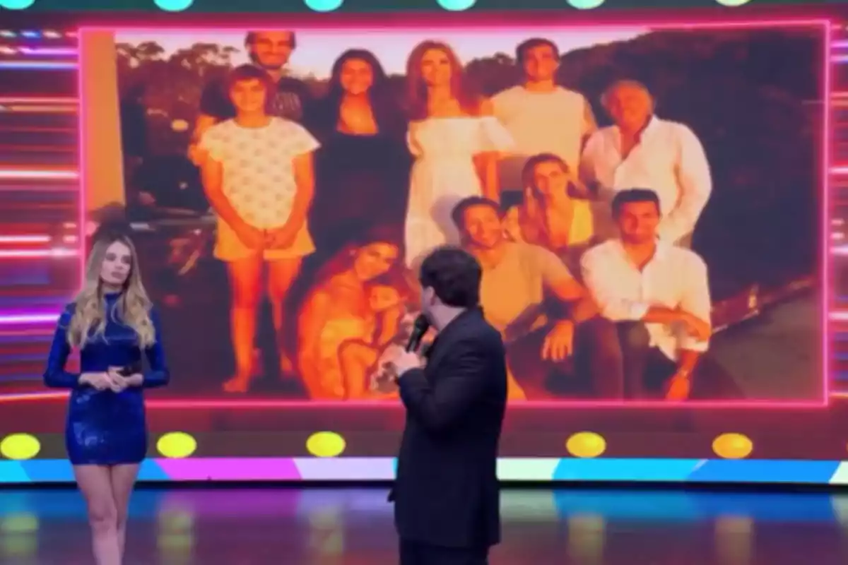 Dos presentadores en un set de televisión observan una gran pantalla que muestra una foto familiar con varias personas posando al aire libre Dos presentadores en un set de televisión observan una gran pantalla que muestra una foto familiar con varias personas posando al aire libre