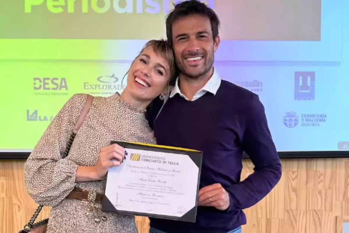 Dos personas sonríen mientras una de ellas sostiene un diploma frente a una pantalla con logotipos de fondo Dos personas sonríen mientras una de ellas sostiene un diploma frente a una pantalla con logotipos de fondo