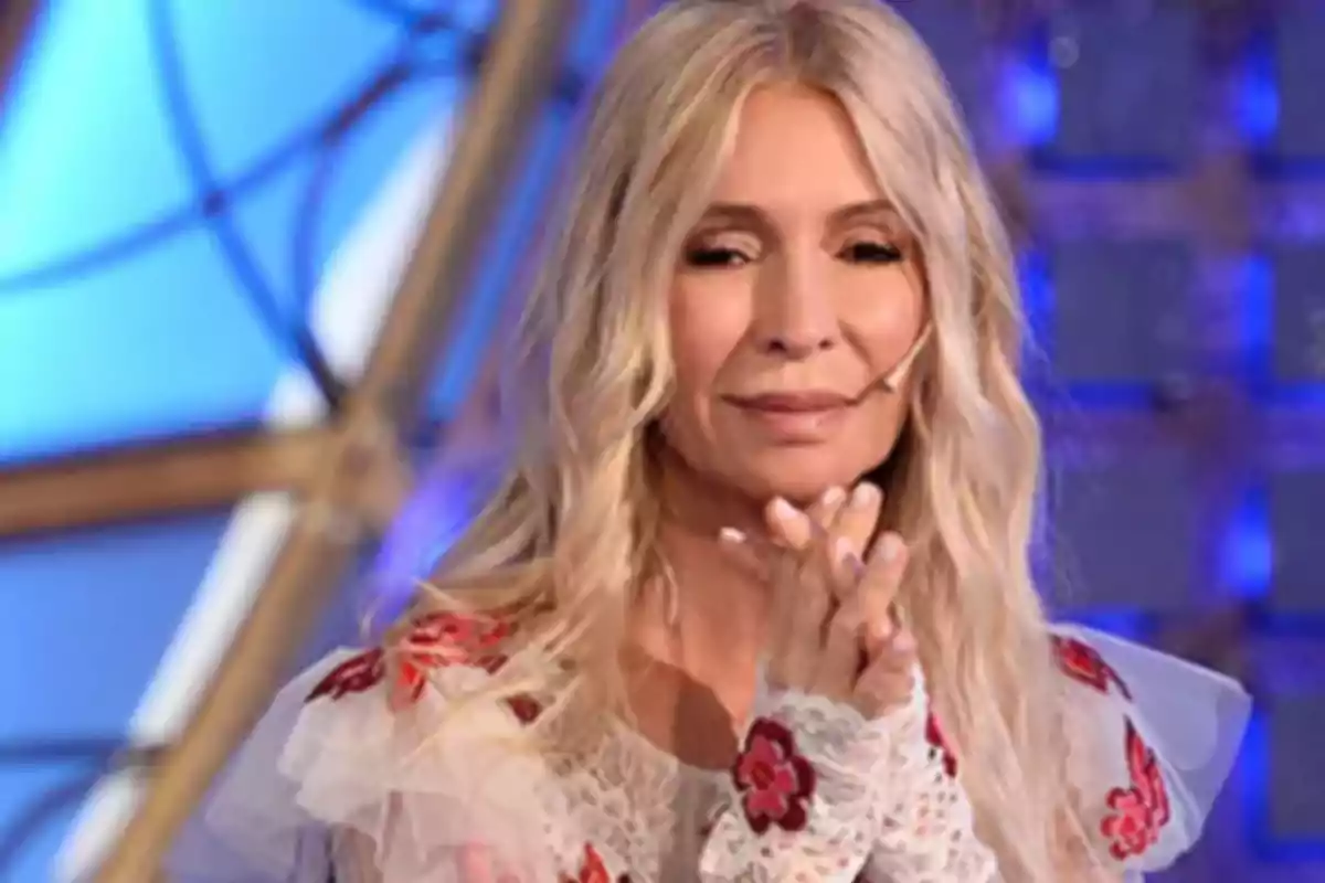 Blonde woman with long, loose hair, wearing a headset microphone and a white dress with red details, on a brightly lit television set