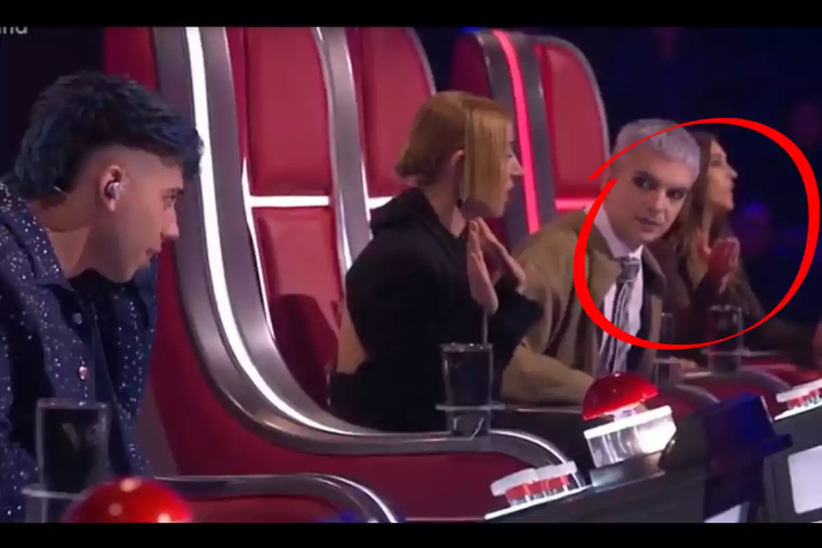 Cuatro personas sentadas en sillas rojas de un programa de talentos, una de ellas está rodeada por un círculo rojo y mira hacia un lado