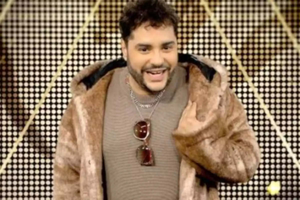 Hombre sonriente con abrigo de peluche marrón y gafas colgadas al cuello frente a un fondo de luces circulares Hombre sonriente con abrigo de peluche marrón y gafas colgadas al cuello frente a un fondo de luces circulares
