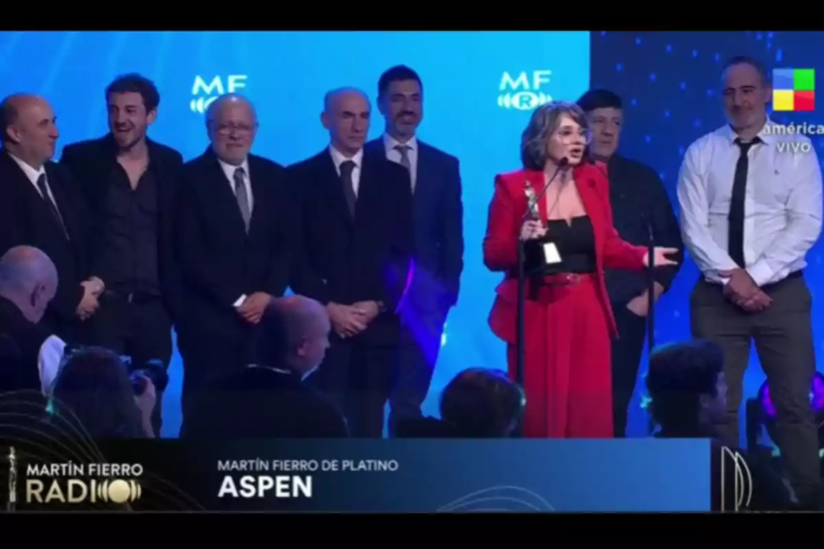 Un grupo de personas en el escenario durante una premiación, una mujer con traje rojo sostiene un trofeo y habla al micrófono mientras los demás la acompañan, el fondo es azul y se observa el logo de un canal de televisión en la esquina. Un grupo de personas en el escenario durante una premiación, una mujer con traje rojo sostiene un trofeo y habla al micrófono mientras los demás la acompañan, el fondo es azul y se observa el logo de un canal de televisión en la esquina.