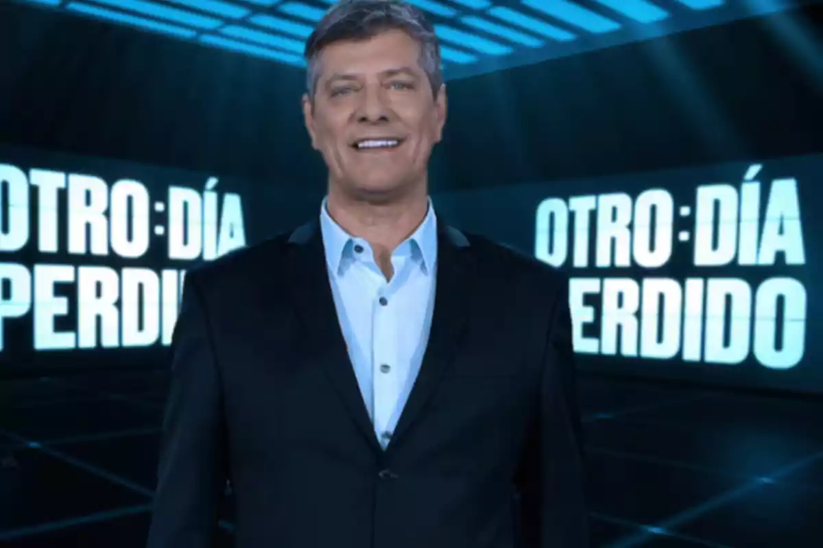 Hombre de cabello canoso y traje oscuro sonríe frente a un fondo con letras grandes que dicen otro día perdido Hombre de cabello canoso y traje oscuro sonríe frente a un fondo con letras grandes que dicen otro día perdido