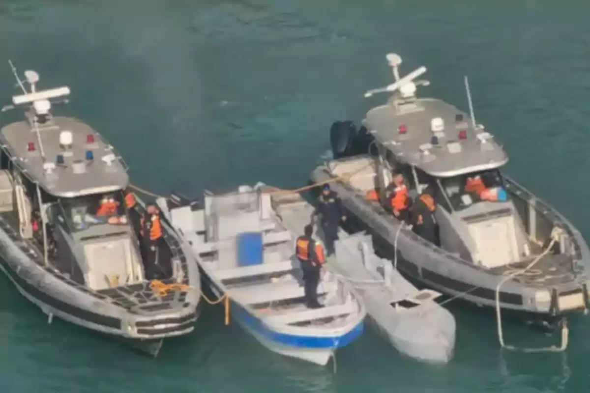 Dos lanchas patrulla rodean y aseguran una embarcación pequeña y un semisumergible en el mar mientras varias personas con chalecos naranjas trabajan en la operación