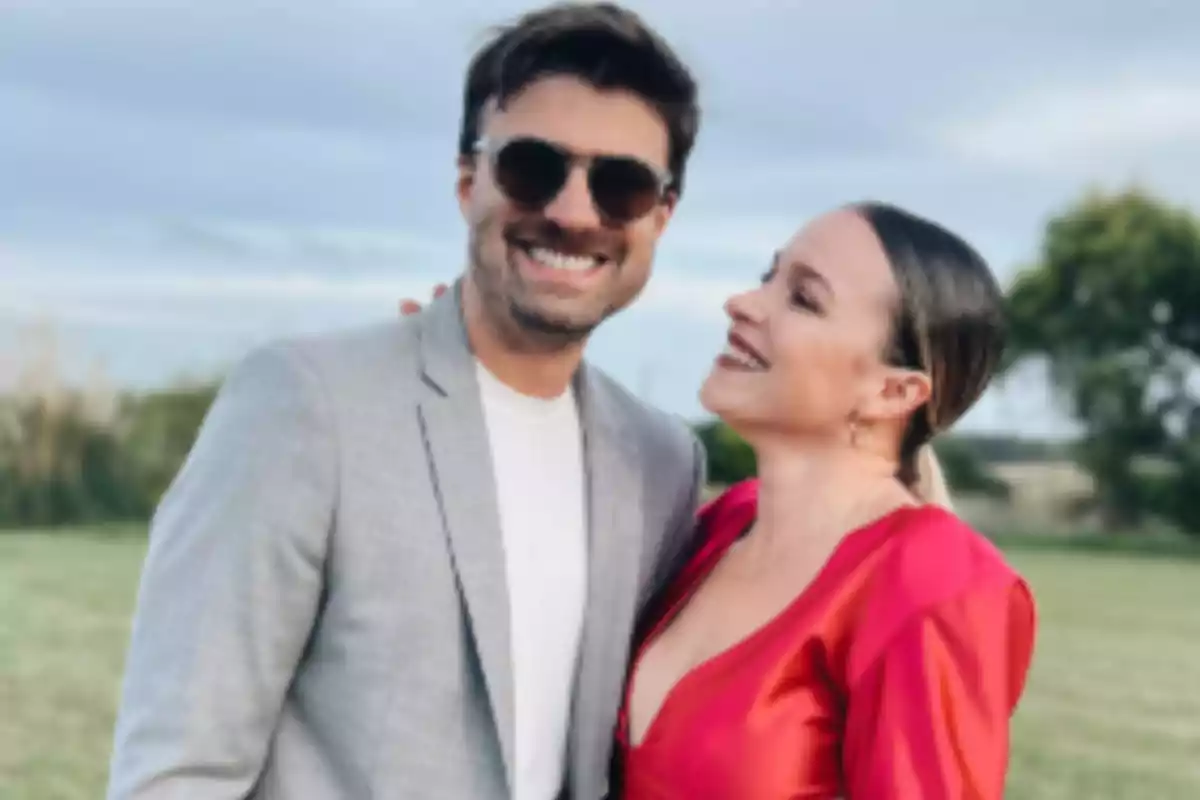 Pareja sonriente posando al aire libre en un entorno natural, él lleva saco gris y gafas de sol, ella viste un vestido rojo brillante