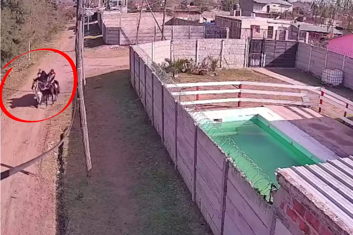 Dos personas montan a caballo por una calle de tierra junto a una casa con piscina y un muro de concreto.