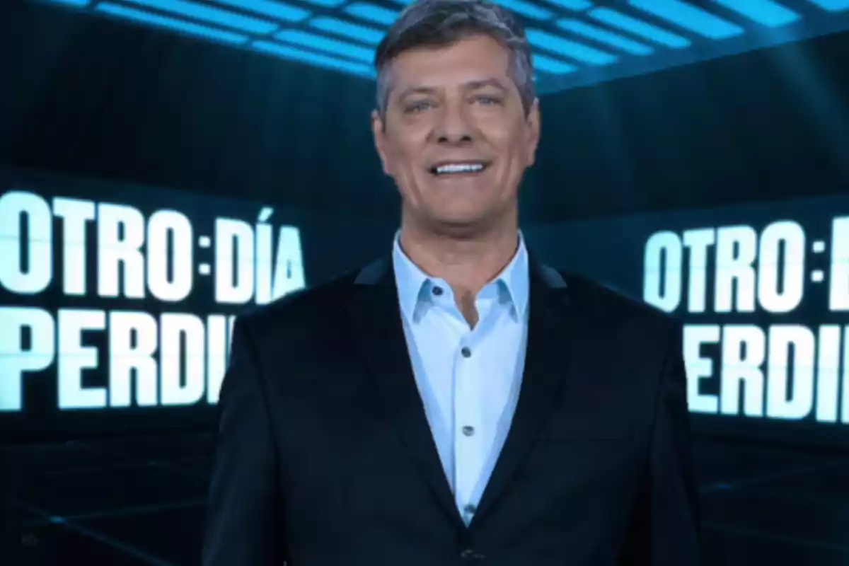 Hombre de cabello canoso y traje oscuro sonríe frente a un fondo con letras grandes y luminosas que dicen "OTRO DÍA PERDIDO"