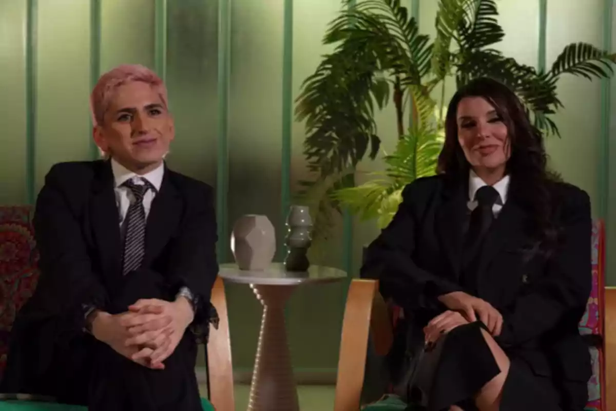 Dos personas sentadas en sillones, ambas con traje oscuro y corbata, una con cabello corto y teñido de rosa y la otra con cabello largo y oscuro, al fondo hay plantas y una mesa pequeña con jarrones decorativos. Dos personas sentadas en sillones, ambas con traje oscuro y corbata, una con cabello corto y teñido de rosa y la otra con cabello largo y oscuro, al fondo hay plantas y una mesa pequeña con jarrones decorativos.