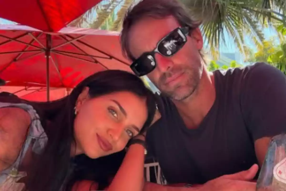 Pareja sentada en una terraza al aire libre con sombrillas rojas y palmeras de fondo