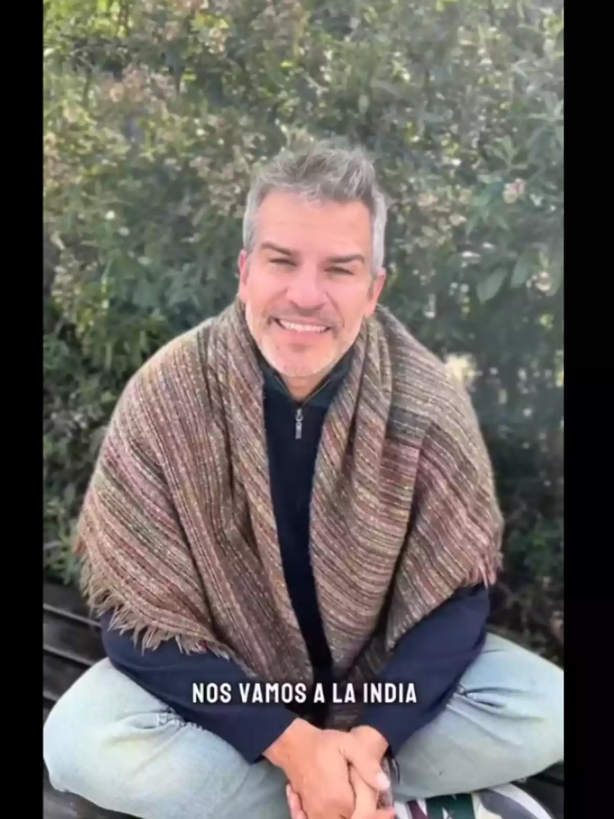 Gray-haired man with a short beard smiles while sitting outdoors with a shawl over his shoulders, and the phrase Gray-haired man with a short beard smiles while sitting outdoors with a shawl over his shoulders, and the phrase