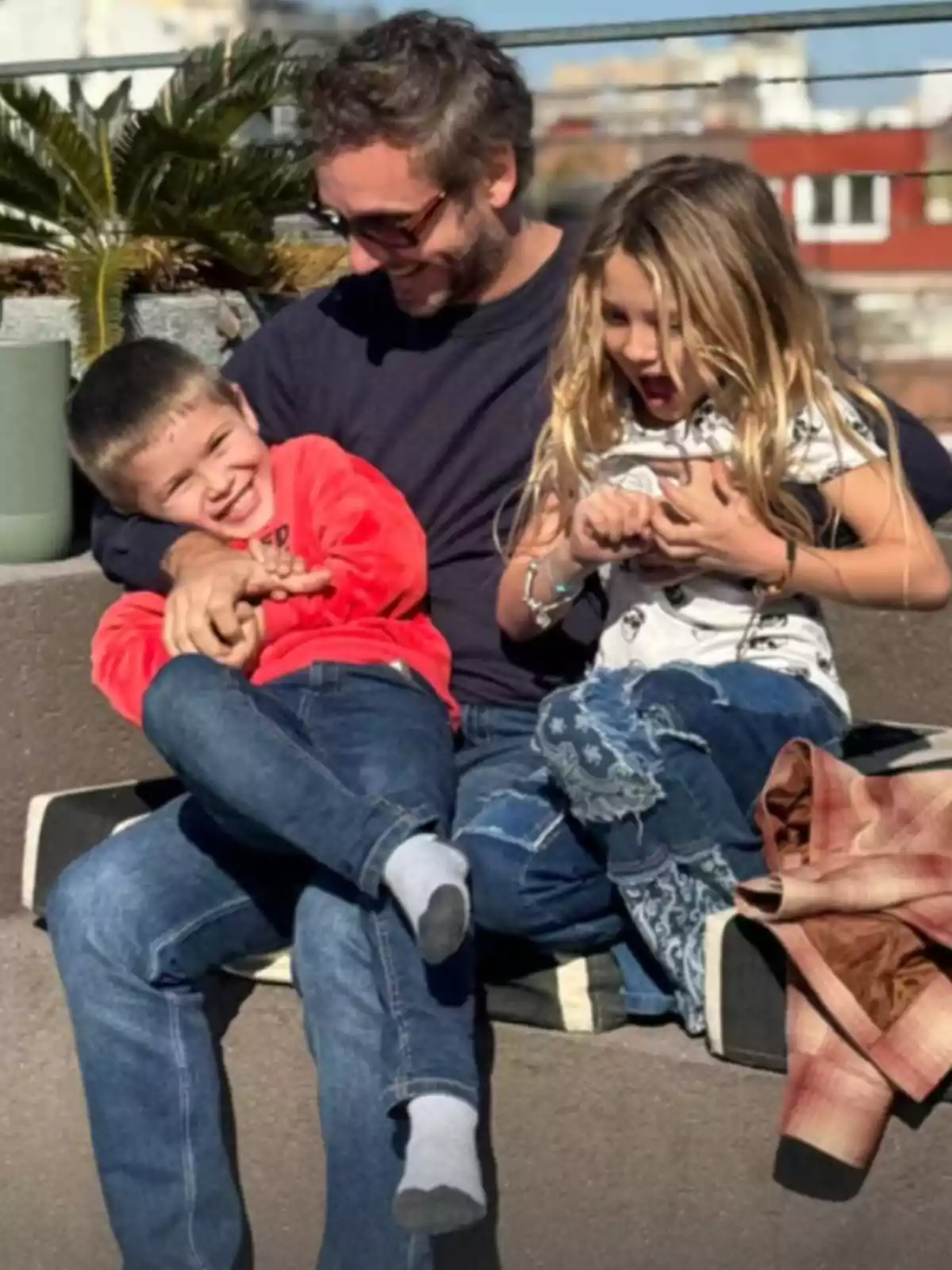 Un hombre sonriente abraza a un niño y una niña mientras se sientan juntos al aire libre disfrutando de un momento alegre bajo el sol Un hombre sonriente abraza a un niño y una niña mientras se sientan juntos al aire libre disfrutando de un momento alegre bajo el sol