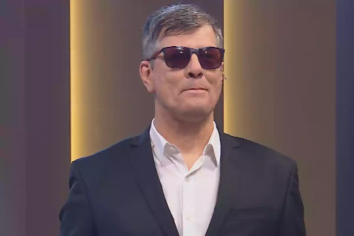 Short-haired, gray-haired man wearing a dark suit, white jersey, and sunglasses in a well-lit indoor setting.