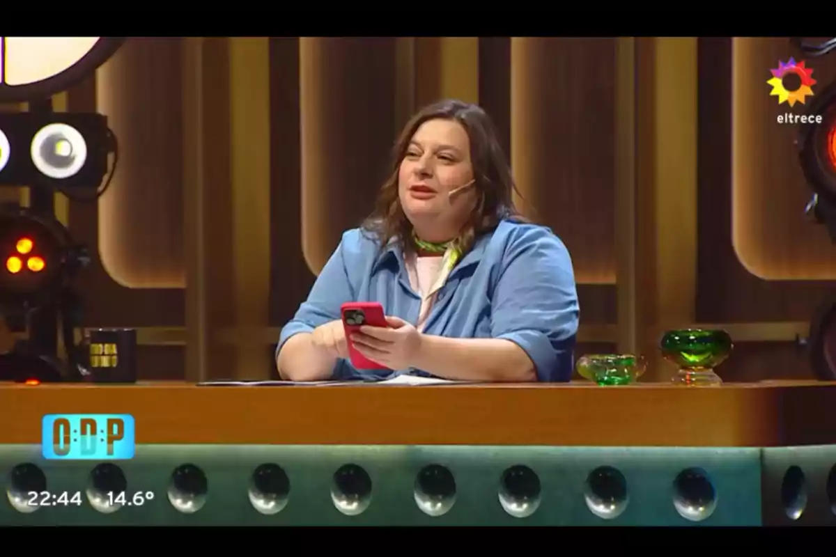 Woman sitting at a TV show desk holding a red cellphone and wearing a headset microphone with a background of lights and modern decor Woman sitting at a TV show desk holding a red cellphone and wearing a headset microphone with a background of lights and modern decor