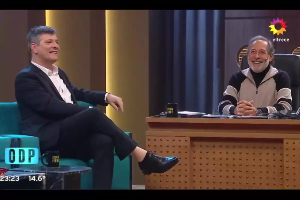 Two men smile and talk on a television set, one sitting in a turquoise armchair and the other behind a desk, with black mugs on the table and a channel logo in the top right corner. Two men smile and talk on a television set, one sitting in a turquoise armchair and the other behind a desk, with black mugs on the table and a channel logo in the top right corner.