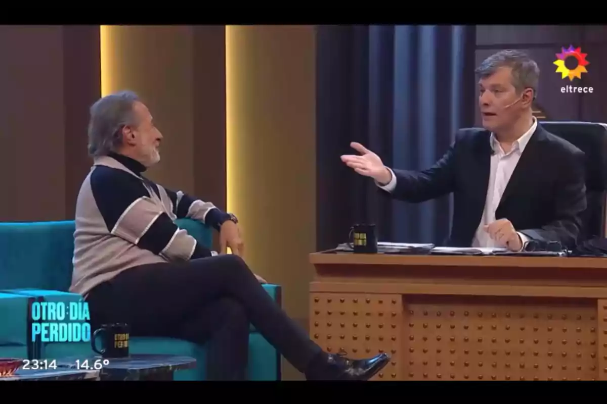 Two men are talking on a television set, one sitting in a blue armchair and the other behind a desk with a black mug while gesturing with his hand. Two men are talking on a television set, one sitting in a blue armchair and the other behind a desk with a black mug while gesturing with his hand.