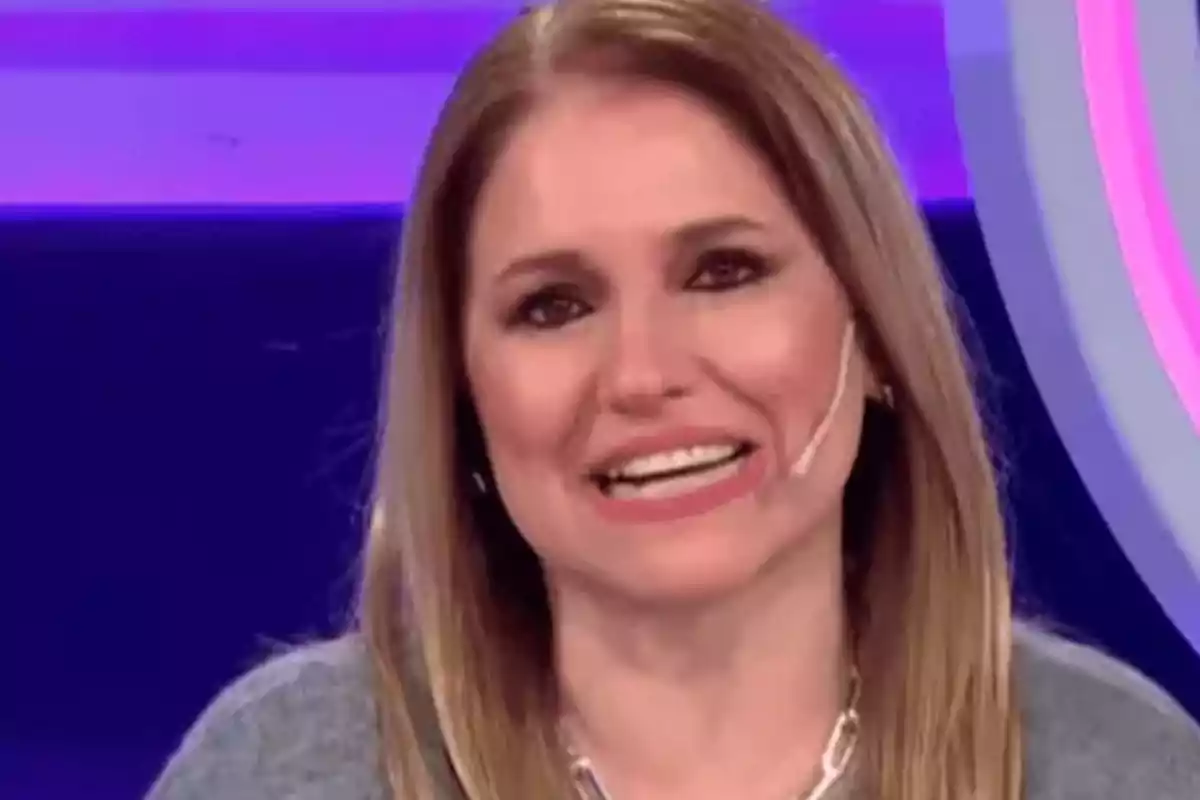 Mujer de cabello rubio lacio sonriendo en un set de televisión con fondo morado y micrófono en la mejilla Mujer de cabello rubio lacio sonriendo en un set de televisión con fondo morado y micrófono en la mejilla
