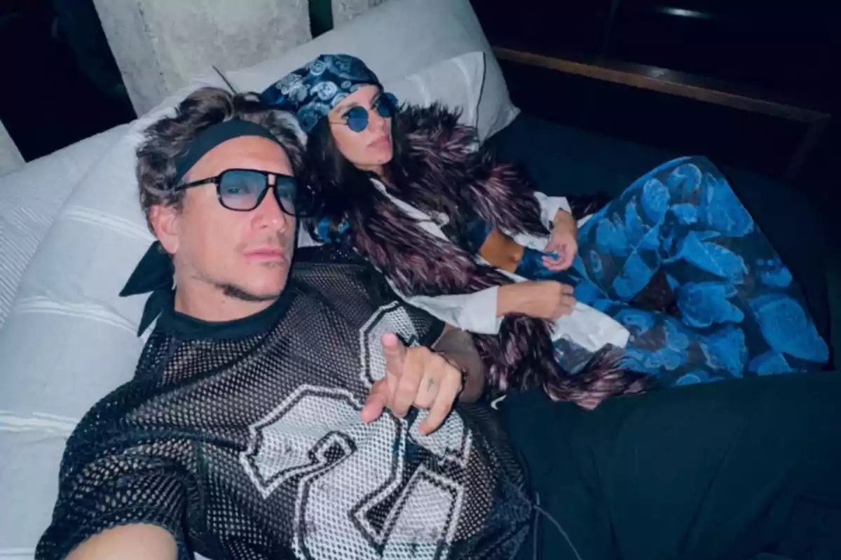 Two people wearing sunglasses and flashy clothes pose reclining on a couch.