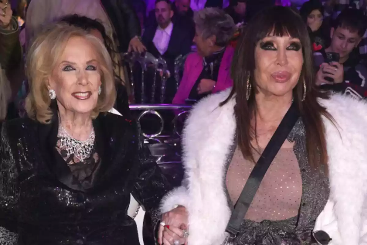Dos mujeres elegantemente vestidas sentadas juntas en un evento nocturno, rodeadas de otras personas en un ambiente festivo Dos mujeres elegantemente vestidas sentadas juntas en un evento nocturno, rodeadas de otras personas en un ambiente festivo