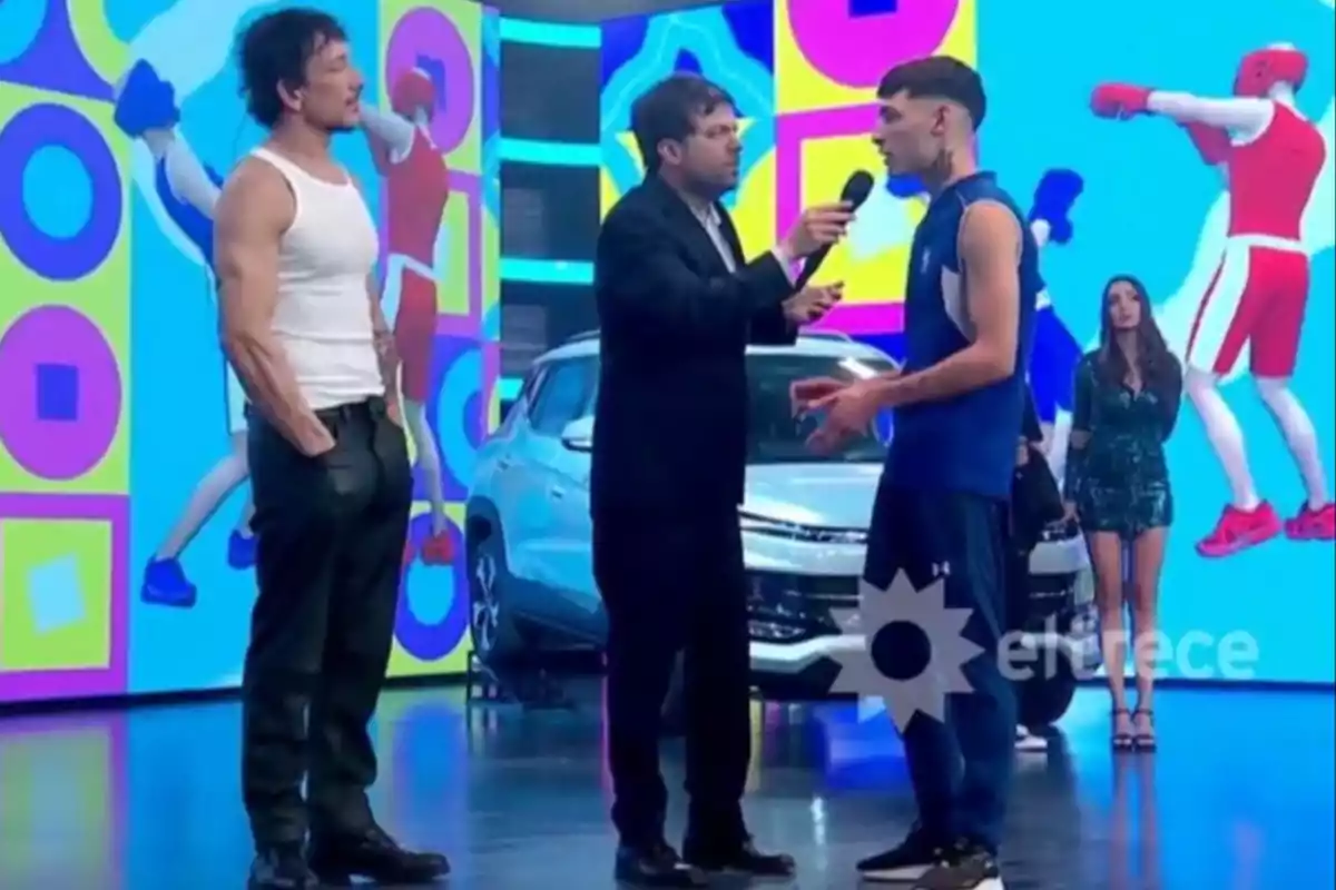Three men are talking on a colorful television set with a white car in the background and a woman standing in the back, while animated boxer figures are shown on the screens. Three men are talking on a colorful television set with a white car in the background and a woman standing in the back, while animated boxer figures are shown on the screens.