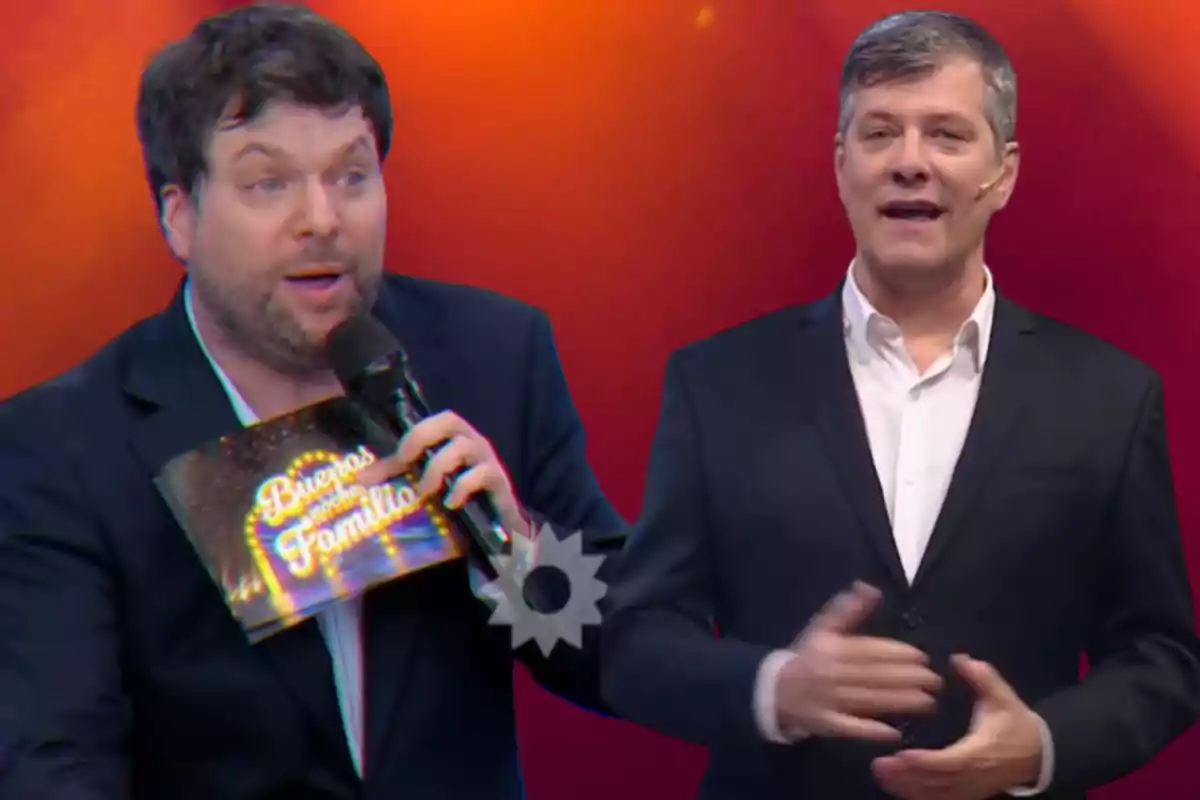Two television hosts dressed in dark suits are on a set with a red background; one is holding a microphone and a folder that says "Good evening, family."