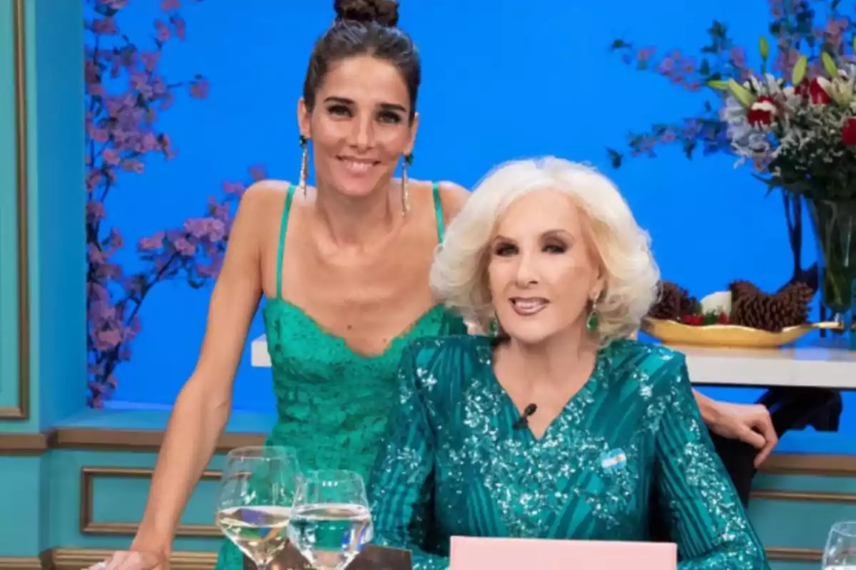 Dos mujeres elegantemente vestidas de verde posan sonrientes en un set de televisión decorado con flores y copas de vino sobre la mesa